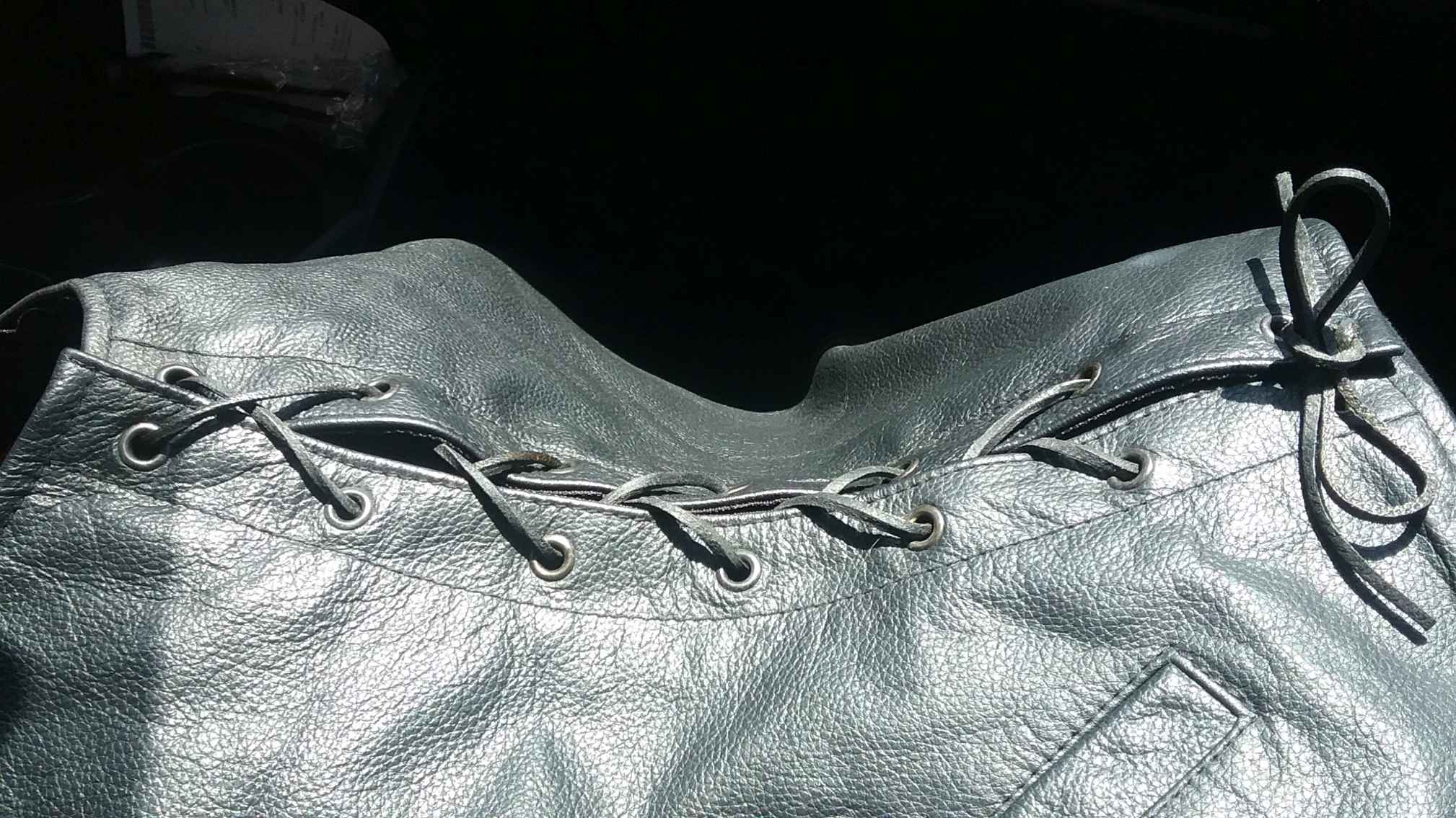 Displayed is the side seam of a black leather vest, joined together with black leather laces tied at one end in a classic bow. Ambient light shines brightly off the leather, giving the impression of a smooth supple finish.