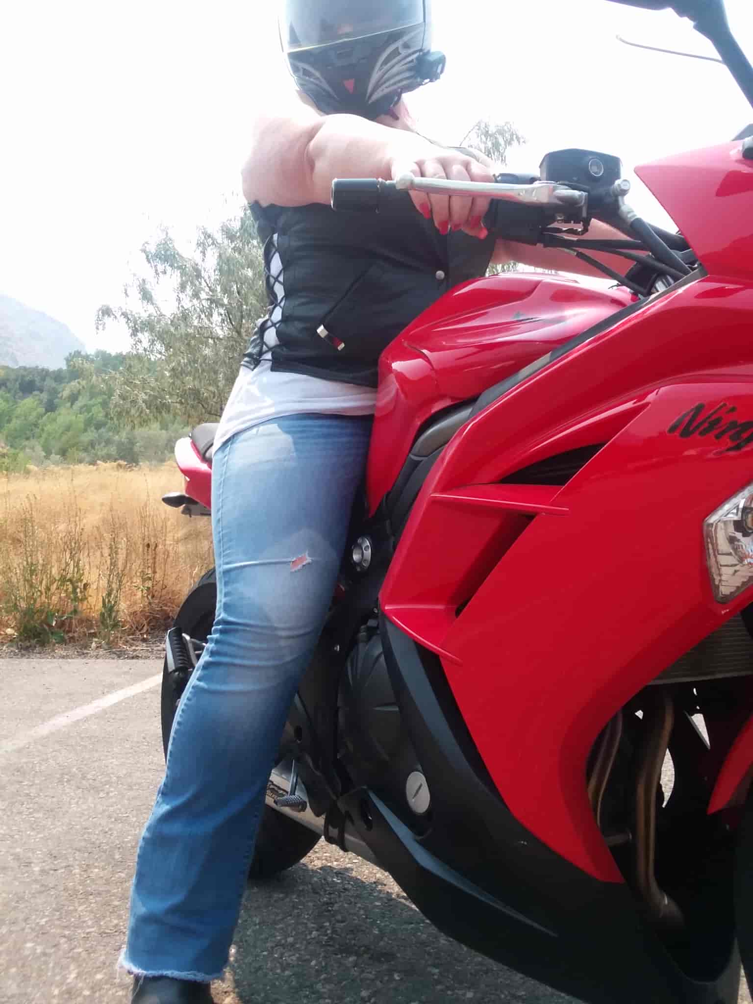 A helmeted woman sits astride a red Kawasaki Ninja 650, looking down at the camera, which faces up from a low vantage. She wears blue jeans, a white tank top and black leather vest. The tank top is visible through laces in the side of the vest. Her right arm is extended and she clutches the throttle of the motorcycle, showing brightly manicured red fingernails.