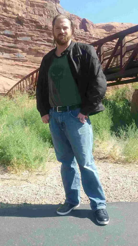A man stands casually in a desert scene, wearing the Shadow Mesh Jacket from Indian Motorcycle. Behind him, a steel bridge spans a wide river basin.