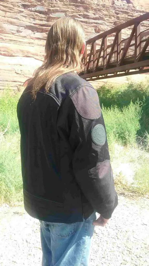 A long-haired man stands quartered away, wearing a dark black jacket with the right arm in the foreground. On the back of the bicep, a round black leather emblem bears the Indian Motorcycle Logo.