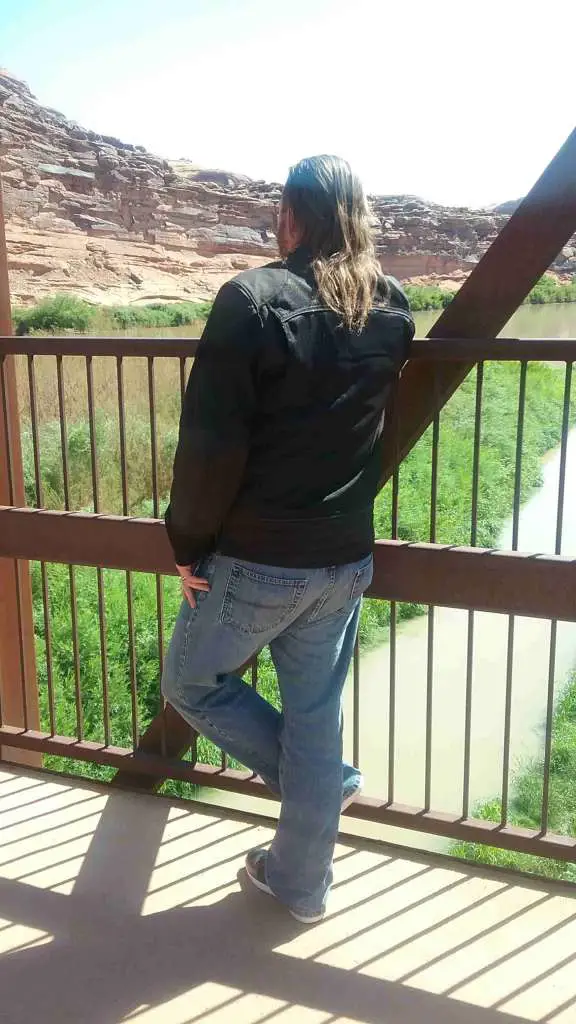 A man stands full-length, quartered away looking out over the guardrail of a rustic steel bridge. He wears the Indian Shadow Mesh jacket, which looks comfortable and slightly menacing in these surroundings.