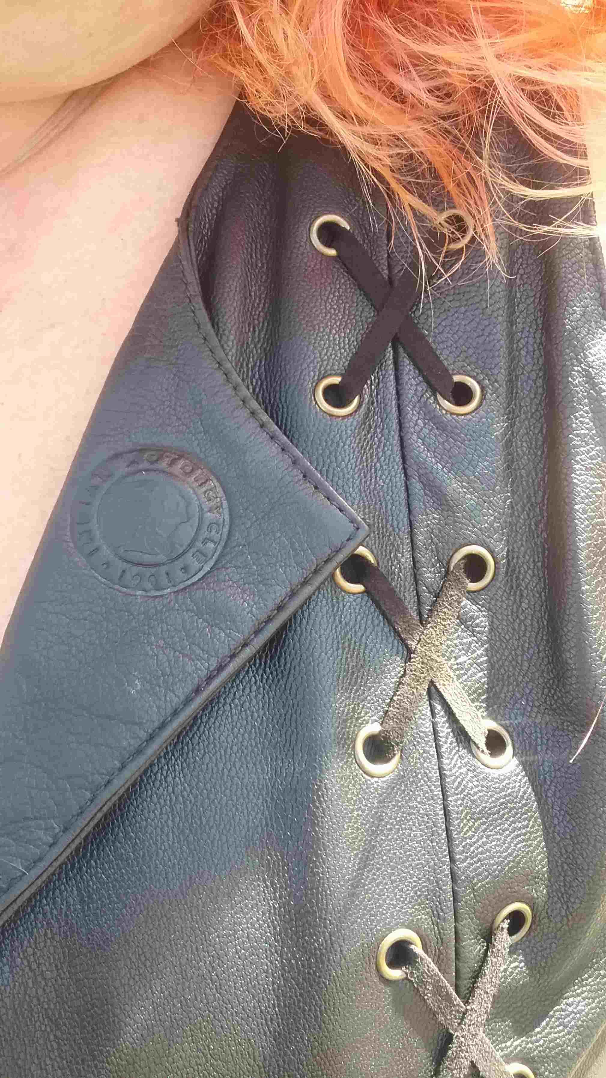 Indian Women's Review, A close up view of criss crossed black leather lacing through antiqued brass eyelets on a leather motorcycle vest, the vest shows a stamped Indian Motorcycle Logo on a trendy leather collar, the woman's red hair flows over the shoulders of the vest.
