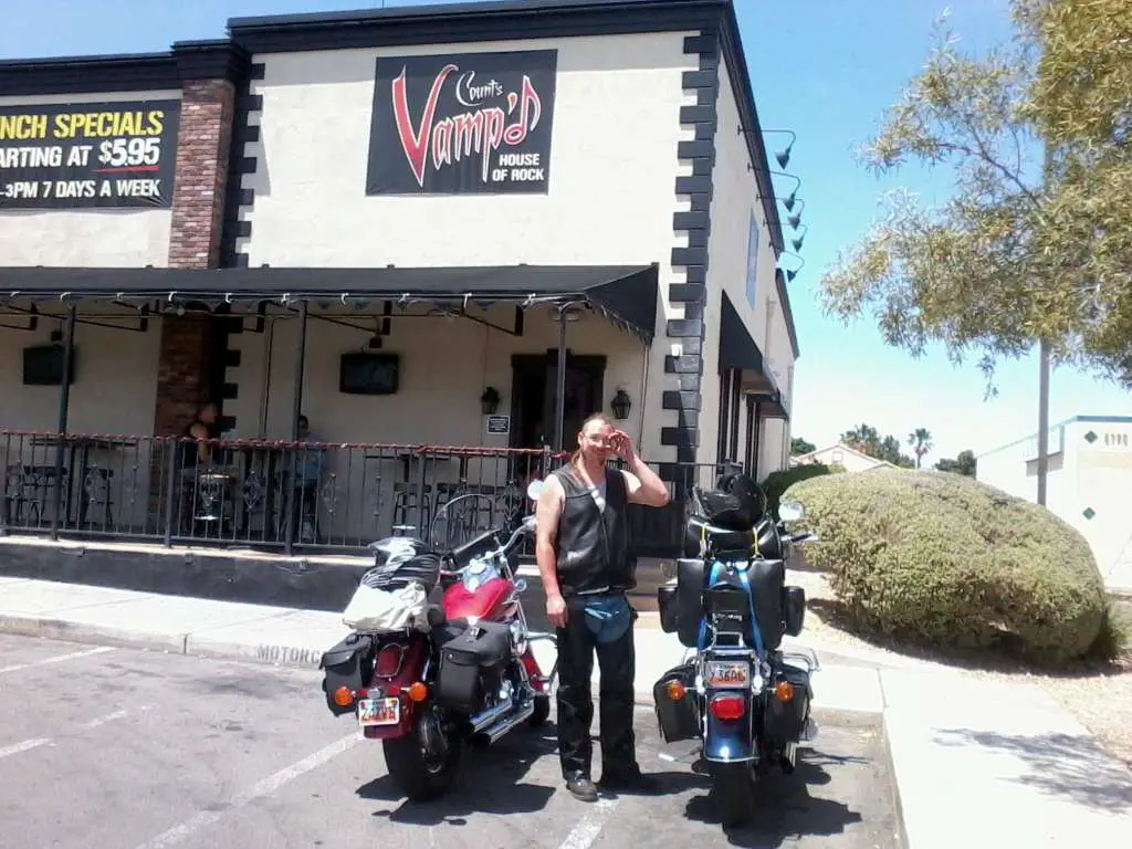 Two luggage loaded cruister motorcycles are parked at a bar. A man wearing a leather vest and chaps shields his face from the Las Vegas sunshine.