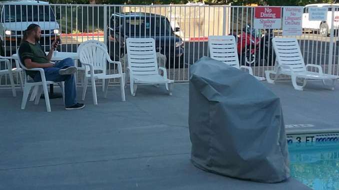 A man checks his phone while drinking coffee in patio chairs poolside in the shade. Two Sport motorcycles are visible in the parking lot behind the pool.