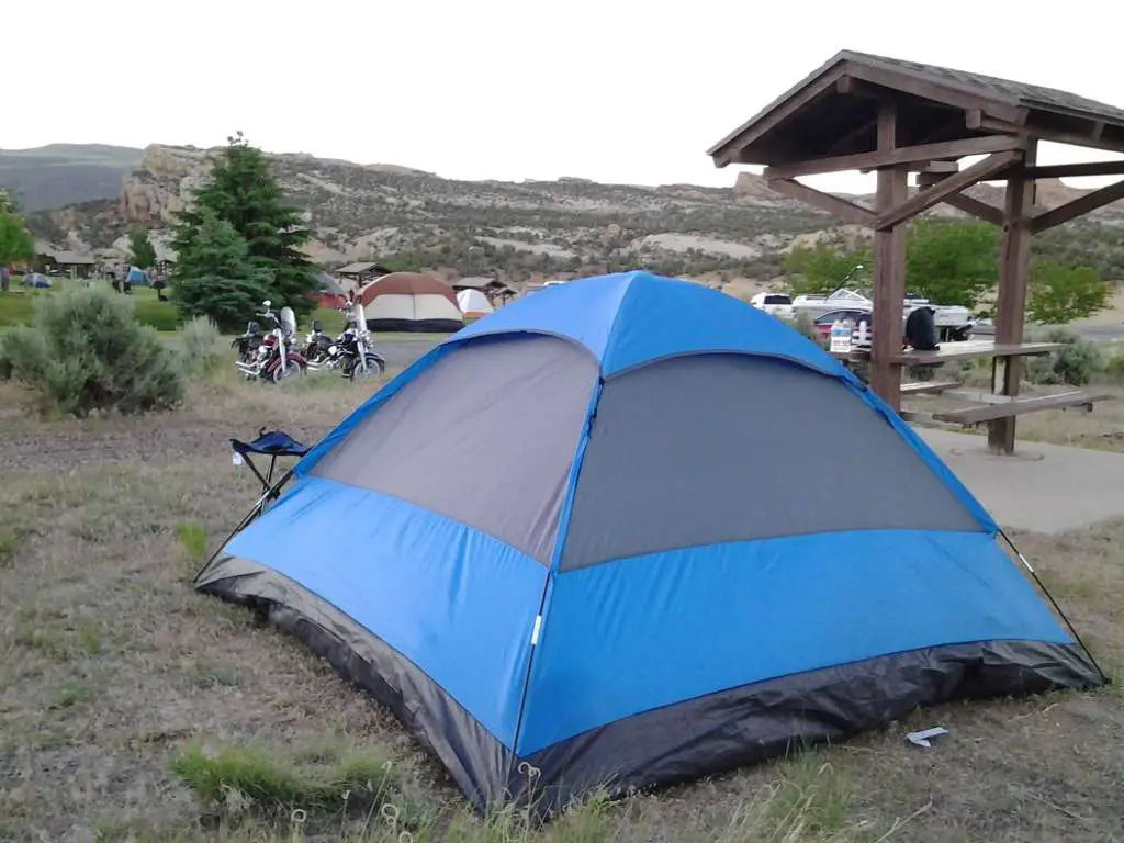 A compact dome tent and tripod chairs are set up in a camp site with beautiful desert scenery, two cruiser style motorcycles are parked in the background.